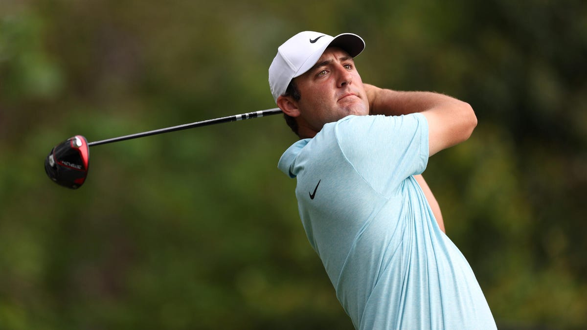 Golfer Scottie Scheffler swinging a club, wearing a light blue shirt and white cap.