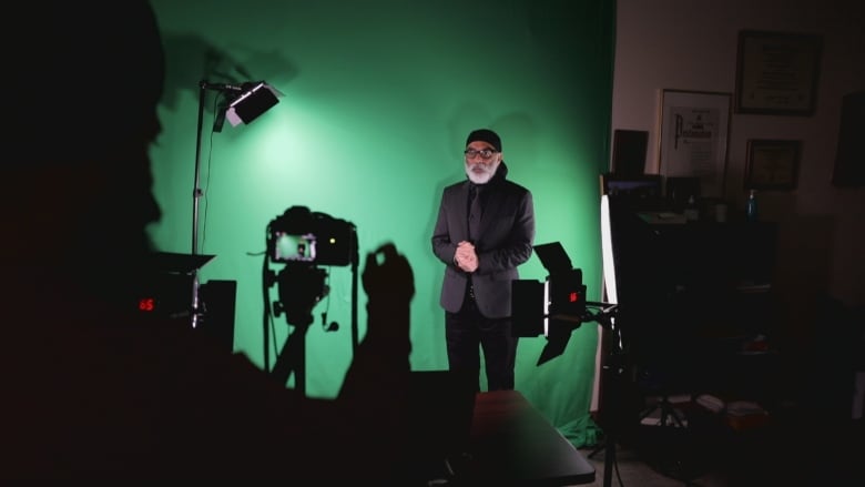 An Indian man wearing a black suit stands in front of a green screen while being video recorded