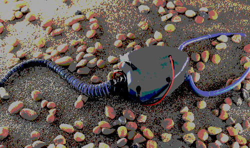 Image of a plastic robot with a triangular body and a long tail, perched on a sandy environment.