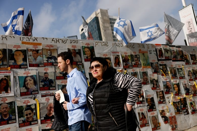 People walk past posters of missing people on a wall with Israeli flags in the background