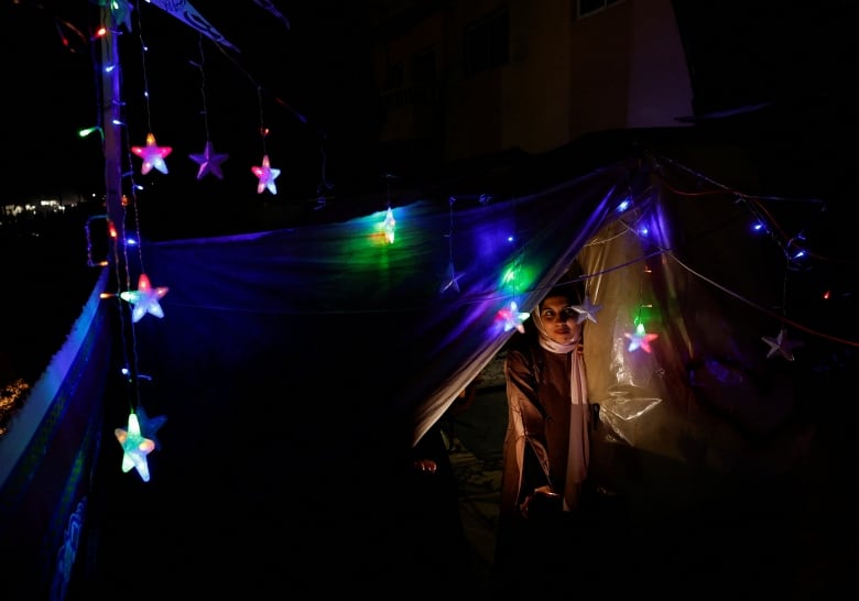 A woman peeks out from a tent at night, surrounded by colourful lights. 