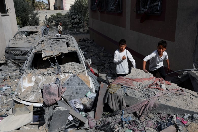 Two boys are shown near bombed out vehicles and rubble in an urban setting.