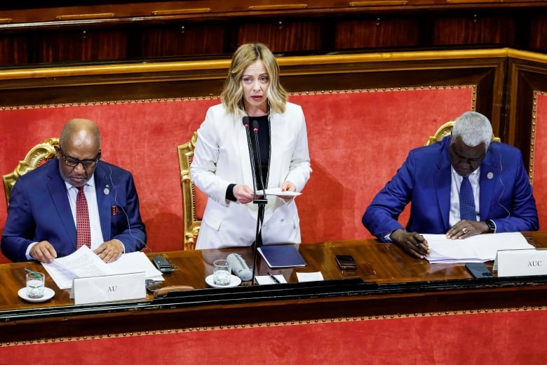 A blonde woman stands between two seated men in blue suits, all of whom are behind an ornate desk.