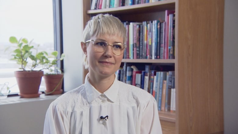 A person wearing a white shirt and with short blonde hair sits in an office.