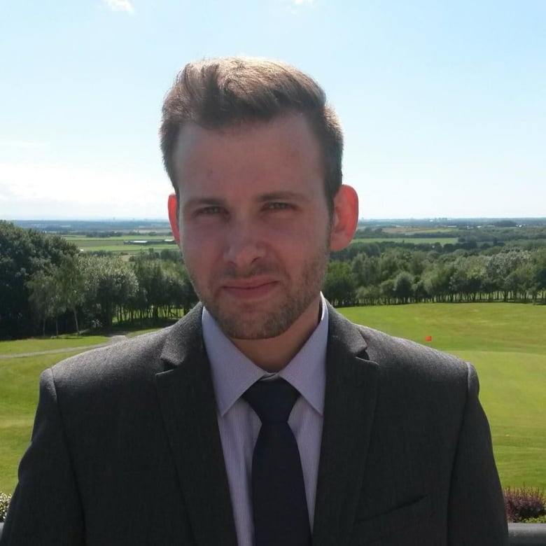 A man in a suit smiles against the backdrop of a sprawling green countryside.