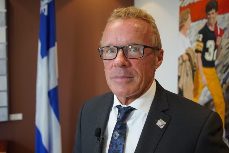 Man in suit and tie stands in front of flag of Quebec in an office.