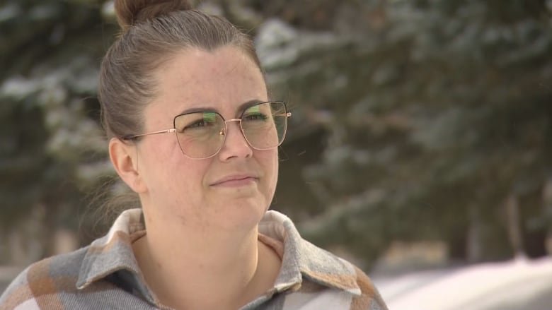 A woman wearing glasses can be seen against a backdrop of trees.