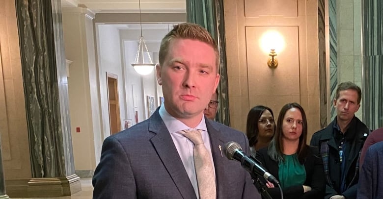 A man in a grey suit and a gold tie stands in front of a microphone. Over his right shoulder a woman in a green shirt and a black sweater crosses her arms. 