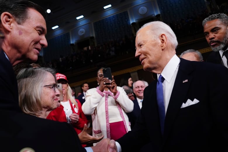 Joe Biden, on the right, shakes hands with a woman in a crowd of people inside a Capitol building chamber. 