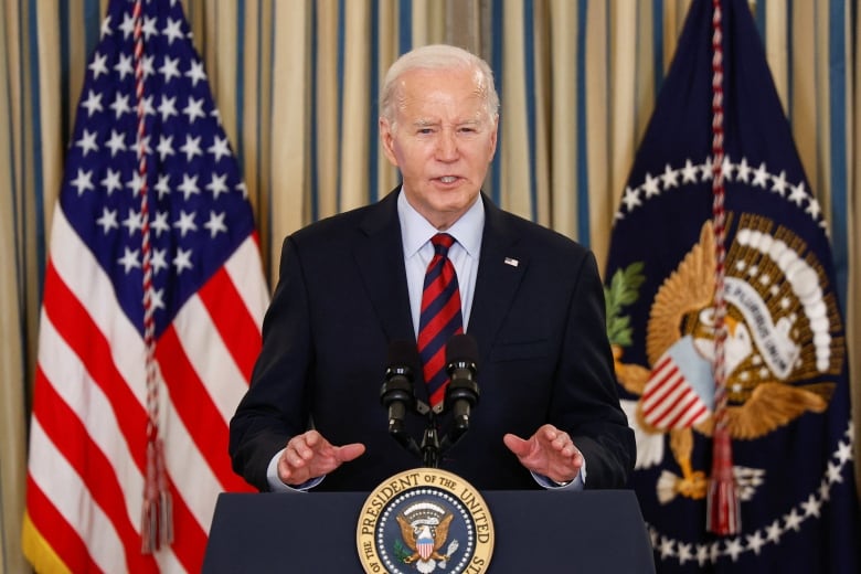 President Joe Biden speaks at a podium, with his hands slightly raised, in front of two flags hanging behind him. 