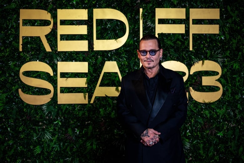 A man with sunglasses on his face and his hair tied back in a ponytail stands in front of a wall covered in green leaves and large gold letters reading "Red Sea FF 2023."