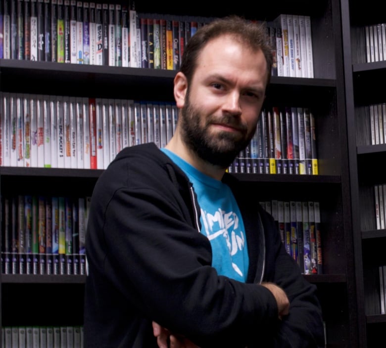 An adult caucasian man with a blue shirt and dark navy hoodie stands in front of a shelf full of video game disc cases.