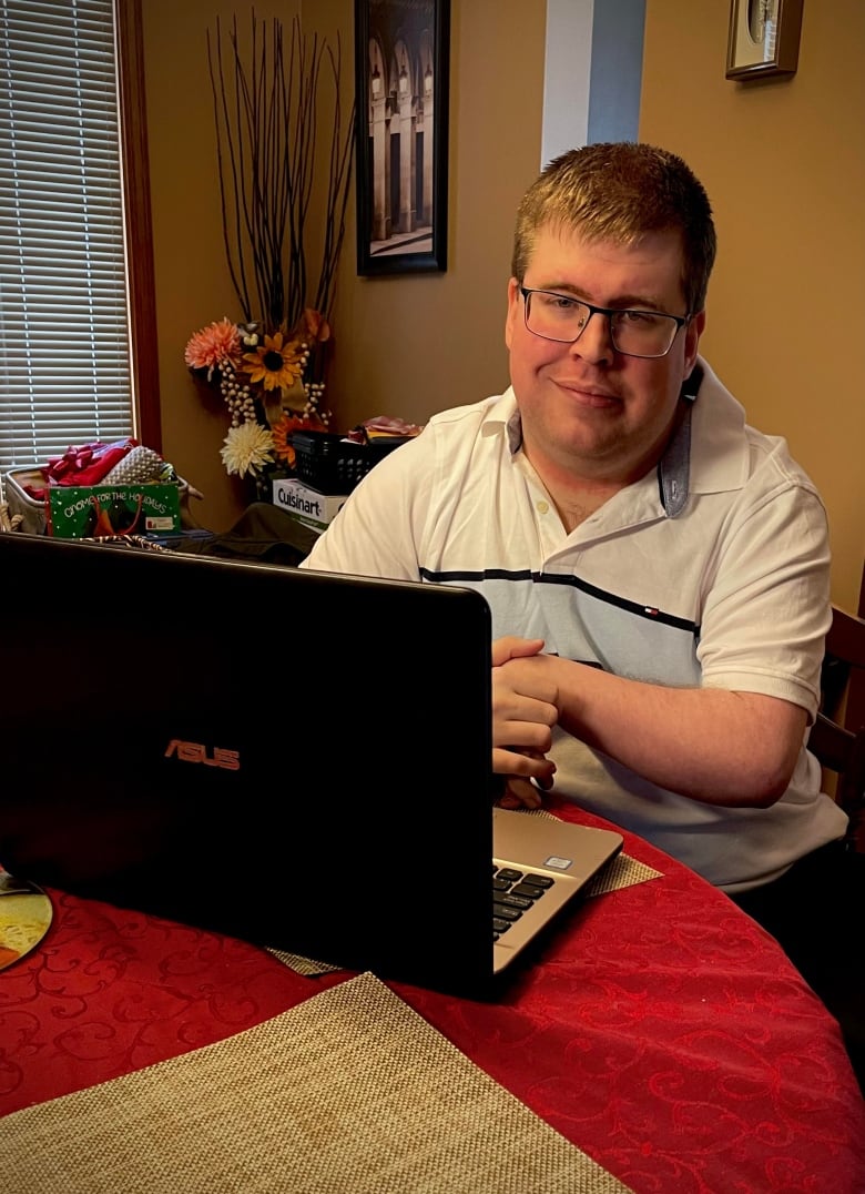 A young man with glasses sits at a table, in front of a laptop.