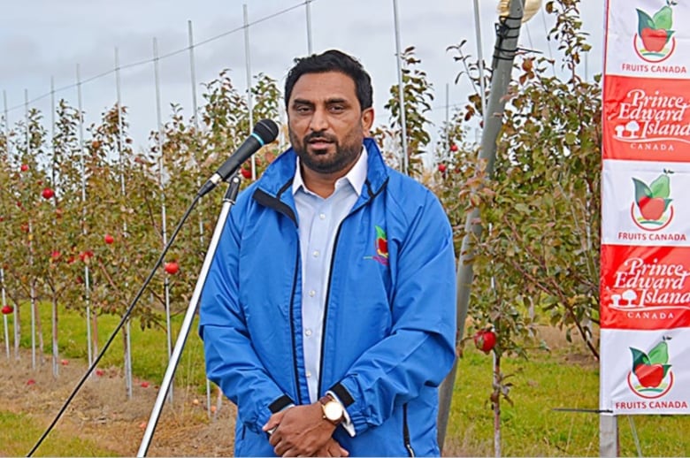 Kamalpreet Khaira is seen speaking into a microphone outside at an orchard and in front of a sign that says P.E.I. and Fruits Canada.