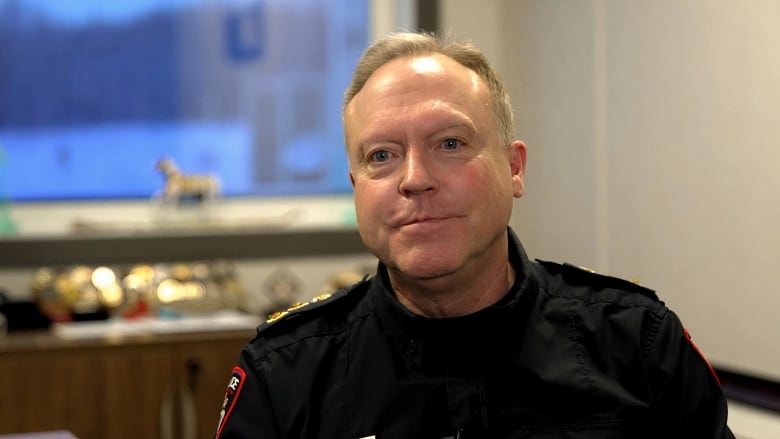 A close-up shot a man smiling at the camera. He has blonde hair and is dressed in a dark police uniform.