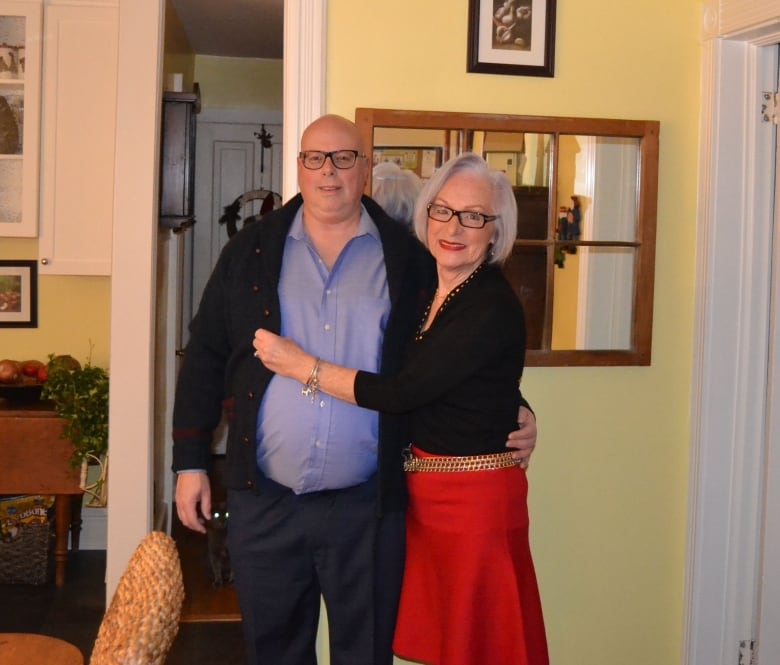 A woman in a red skirt and gold skirt smiles as she embraces a man. They’re standing in a living room. 