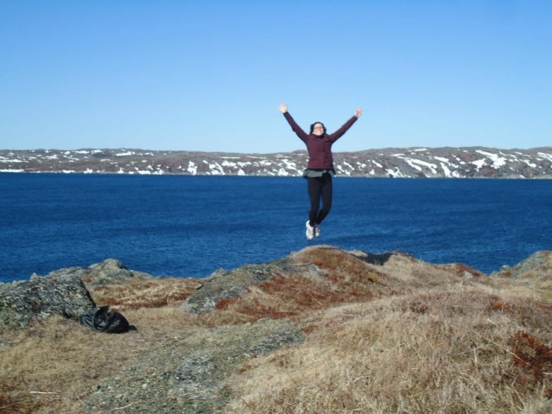 A woman jumps up with her hands in the air. She’s on a grassy patch next to the ocean with snow-covered mountains across the water.  