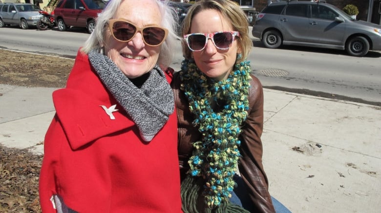 Two smiling women pose for the camera. One of them is wearing a leather jacket while the other wears a bright red jacket with a silver bird broach.