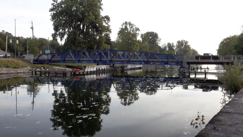 A blue metal bridge is shown over a slow-moving canal, which has algae floating on the surface.