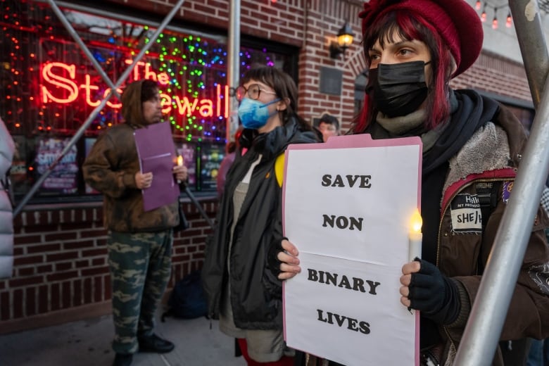 People stand outside a bar, with a red neon-lit sign reading Stonewall Inn in the window. One woman in the foreground, wearing a face mask and a burgundy hat that matches a streak in her dark hair, hold s sign reading "Save non binary lives."