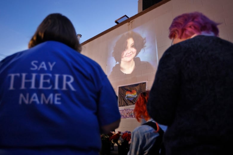 A person, on the left, sits outside in the evening light, wearing a blue jacket with the words "say their name" in white letters on the back, next to another person with short, purple hair. between them is the image of non-binary teenager Nex Benedict on a large screen. 