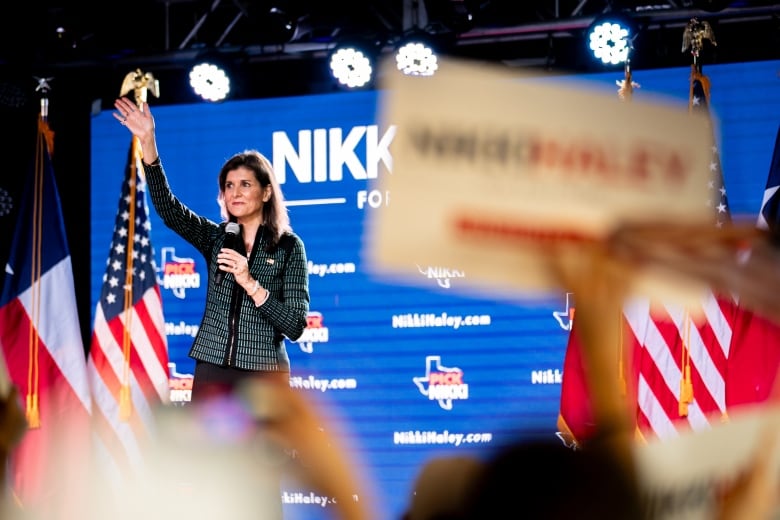 A woman stands on a stage, waving her right hand to a crowd of people holding signs.  