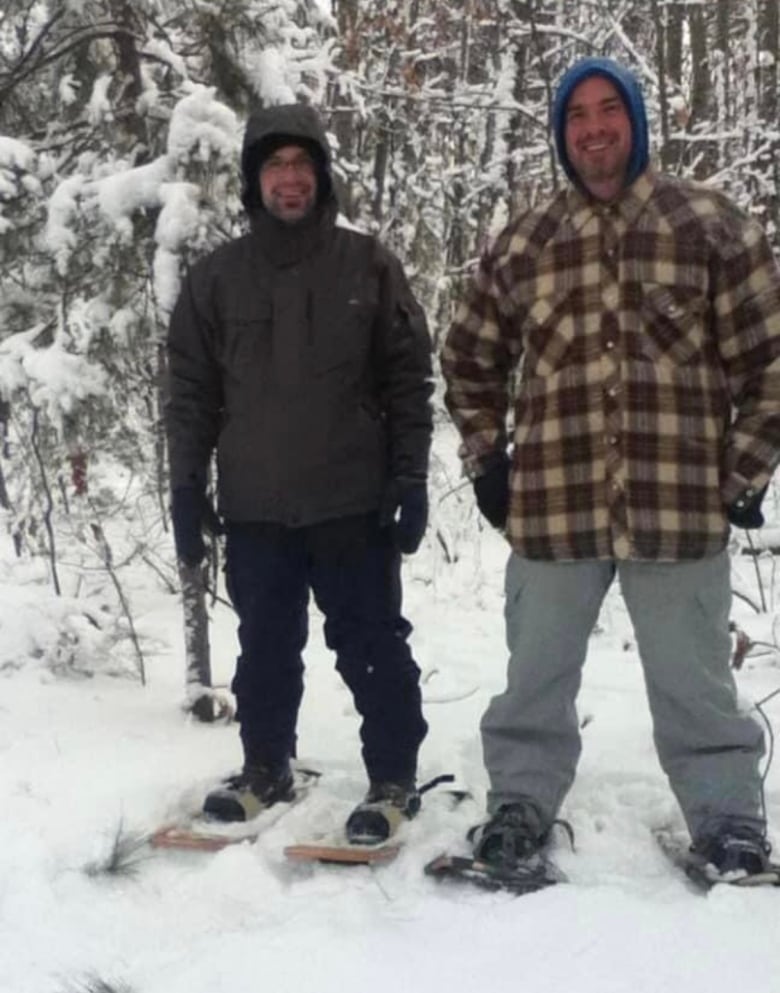 Two smiling men stand next to each other. They’re wearing snowshoes and in a wintry forest. 