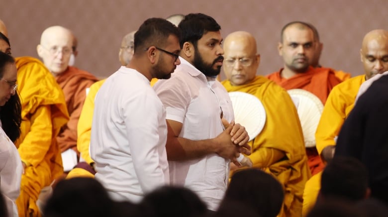 Two men walk by a group of Buddhist monks. The men are wearing white shirts, and the one on the right has a cast on his ring finger.