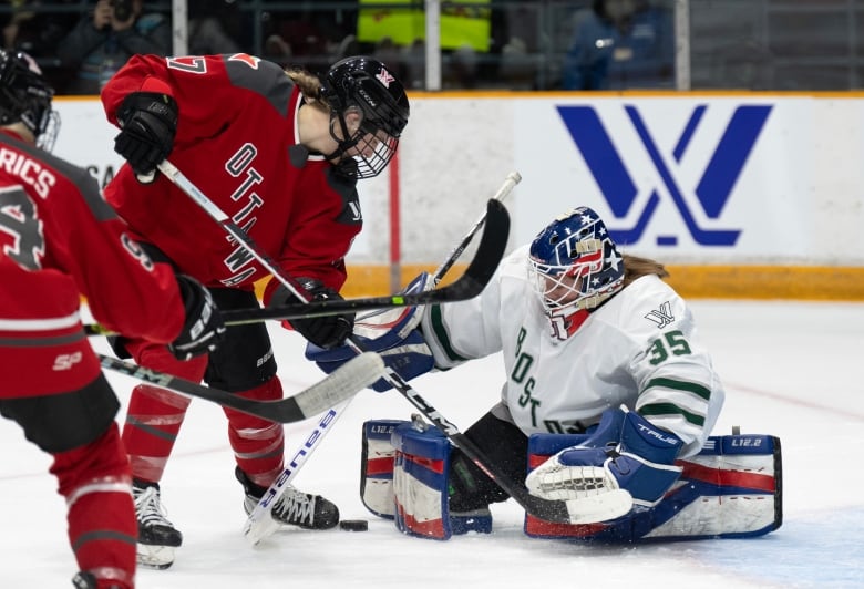 A female ice hockey goaltender makes a save while down on both knees as an opponent digs for a rebound in front of her.