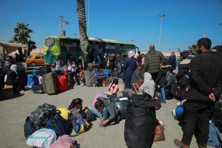 Families with bags and suitcases wait to board a green passenger bus at a depot.