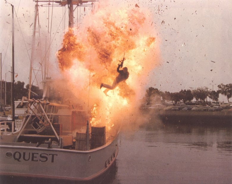 A man is thrown off  a boat with an explosion behind him.