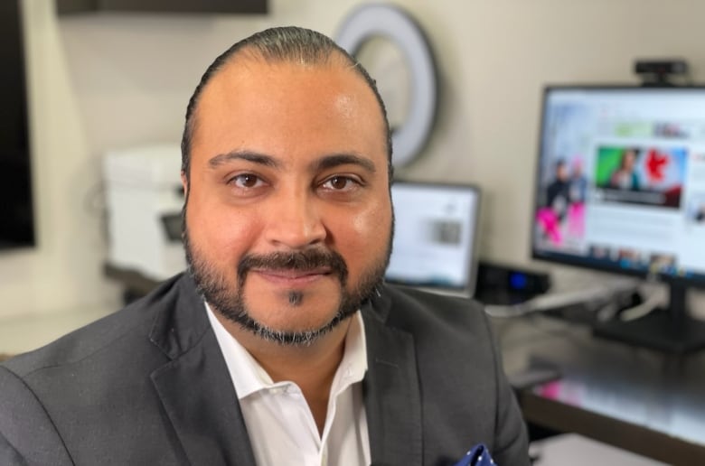 A balding man wearing a grey blazer over a white collared shirt looks directly at the camera with a slight smile on his face. There are two computer monitors in soft focus in the background.