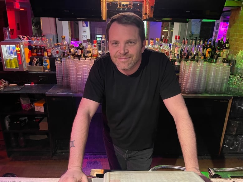 A man stands behind a bar in a nightclub. He looks at the camera and smiles.