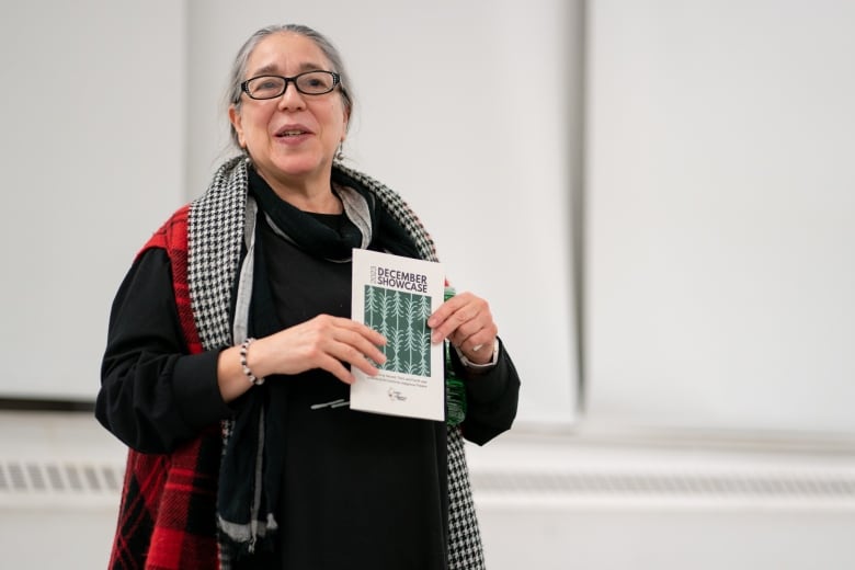 An Indigenous woman standing in a room. She is holding a playbill that says "December Showcase."