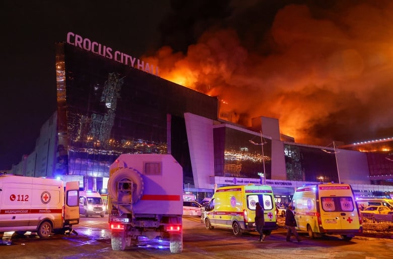 Emergency vehicles parked in front of a large building at night with huge flames and smoke rising above the roof. 