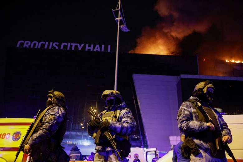 Armed security guards, wearing helmets and fatigues, stand in front of a building with the words "Crocus City Hall" on the top. Flames and smoke are rising above the building. 