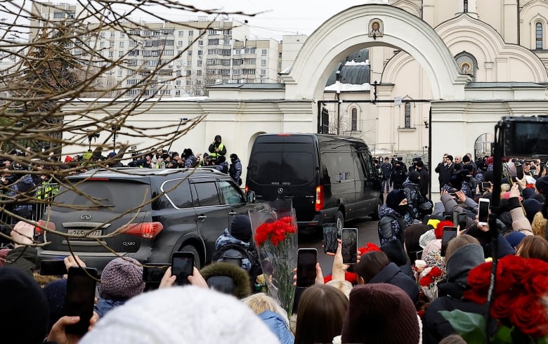 Two black vehicles are shown surrounded by throngs of people near a building.