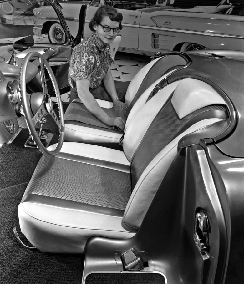 In this black and white archival image, a woman with short hair and black framed glasses sits just inside the interior of a convertible sports car. 