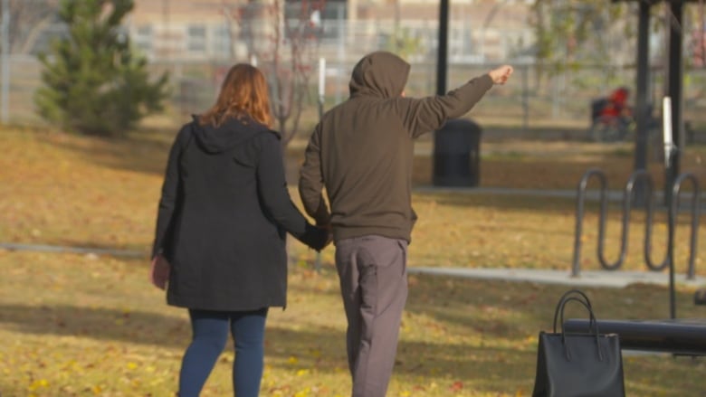 A woman with red hair and a teen in a hoodie walk through a park, holding hands.