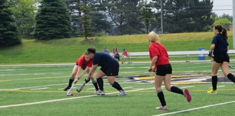 Four female athletes playing field hockey. Two of them are wearing red shirts and the other two are wearing blue shirts.  