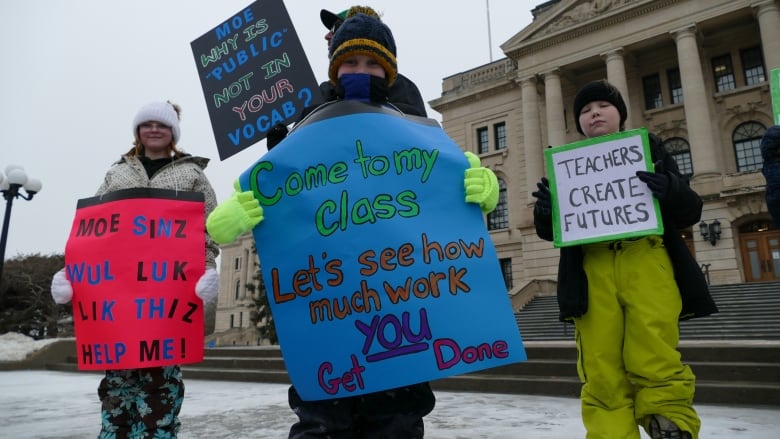 Three children hold signs in support of teachers. A blue sign reads "Come to my class, let's see how much work YOU get done." A white and green sign reads "Teachers create futures." A red sign reads "Moe sinz wul luk lik thiz help me!"