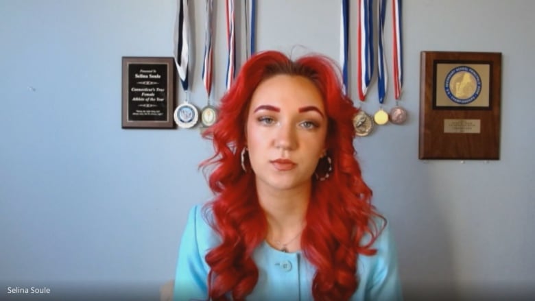 A woman with long red hair sits in front of a wall with several medals and awards.