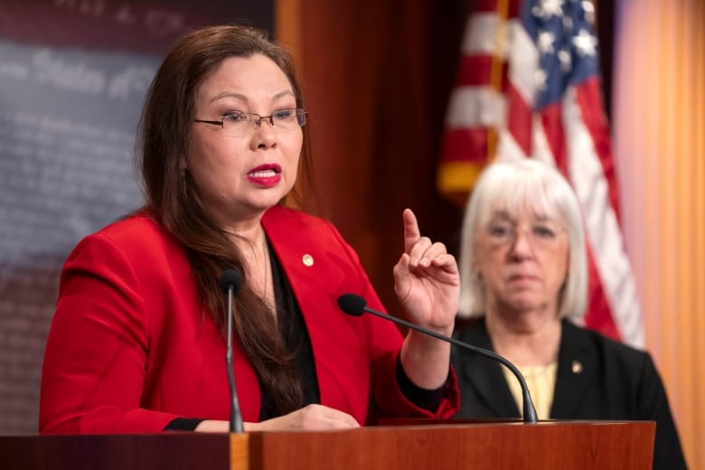 A woman wearing glasses gestures with her hand while speaking into a microphone at a podium. Another woman looks on while on stage with her.