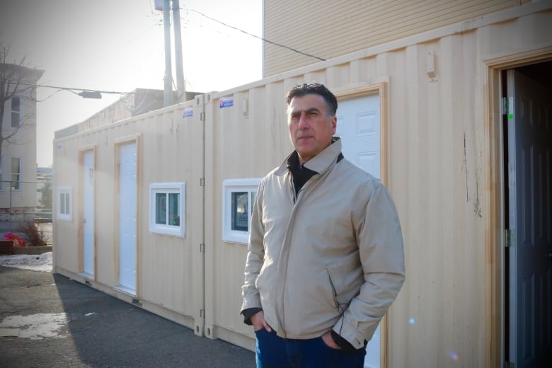 A man in a beige jacket stands in front of a row of beige shipping containers. 