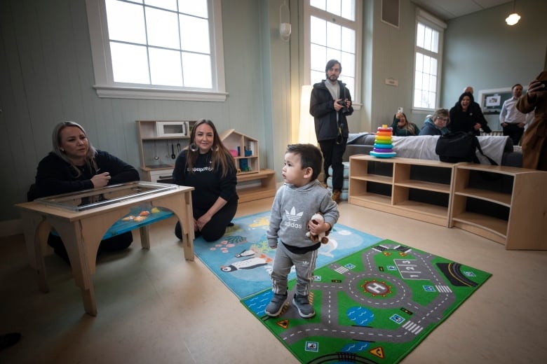 A toddler boy stands in the middle of a wide room with teachers encouraging him from the floor beside him.