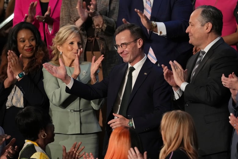 A man wearing a black suit and glasses gestures with his right hand as he stands next to a woman wearing a green suit, clapping her hands, in a crowd of other people applauding. 