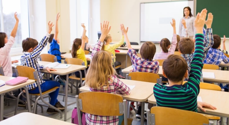 A female teacher stands at the front of a classroom as several students seated at their desks raise their hand.