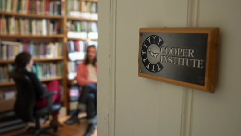 The sign for Cooper Institute is shown on a door, with two staff members sitting in the background out of focus.