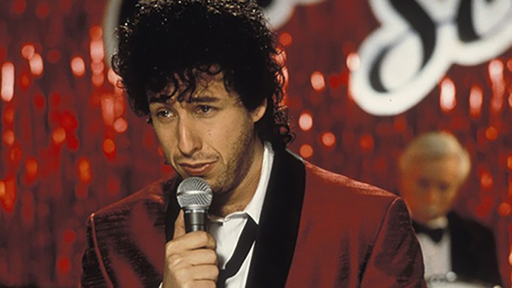 Adam Sandler with curly hair holding a mic and singing in a scene from The Wedding Singer.