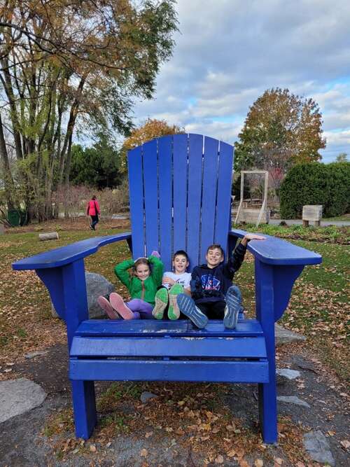 A girl and two boys sit on an oversized blue chair in a park.